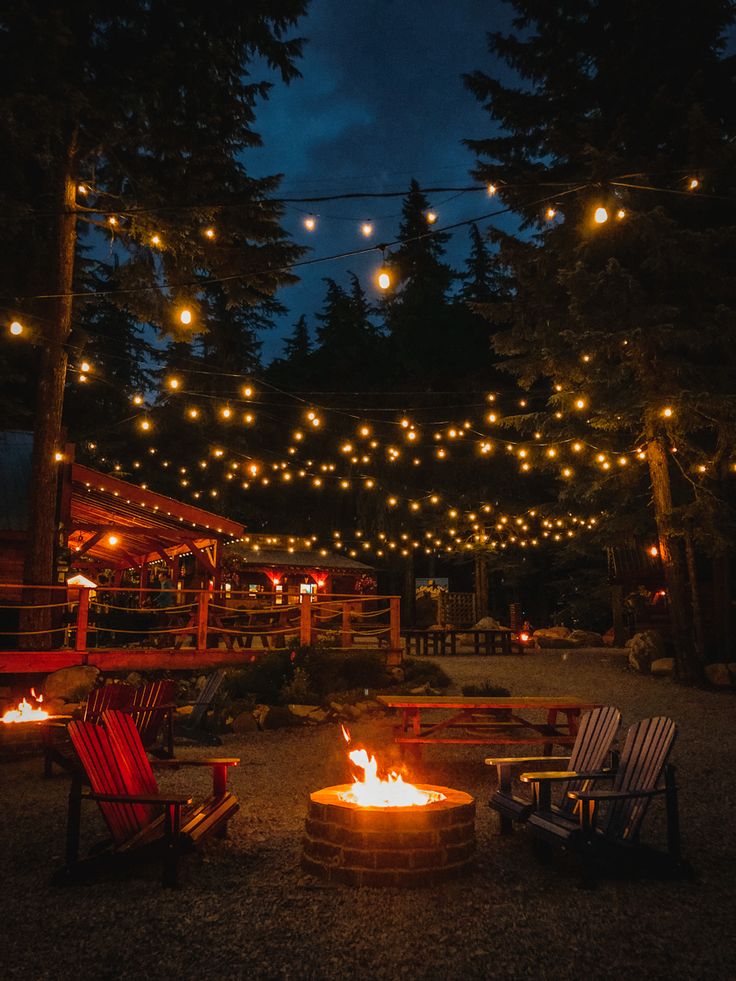 a fire pit surrounded by lawn chairs and string lights at night with trees in the background