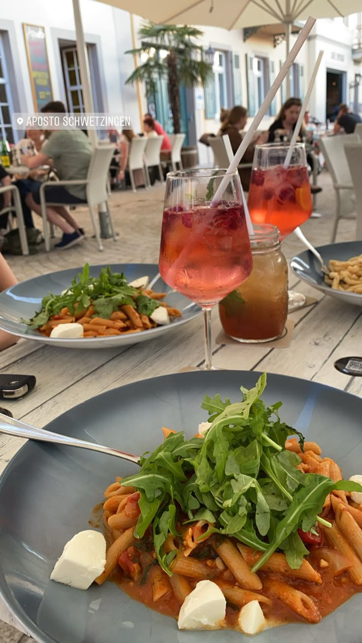 two plates of pasta and vegetables on a table with people sitting at tables in the background