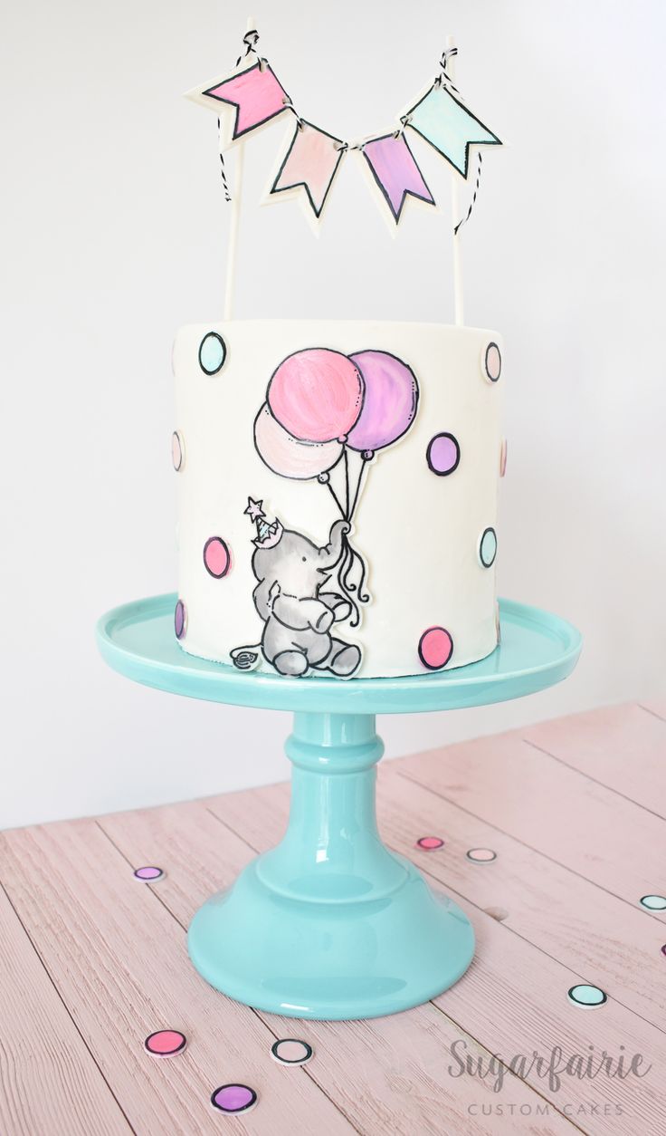 a white cake with pink and blue balloons on top, sitting on a turquoise pedestal