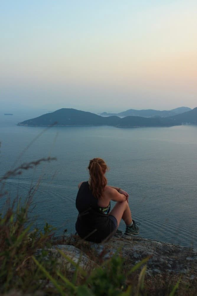 a woman sitting on the edge of a cliff looking out at water and hills in the distance