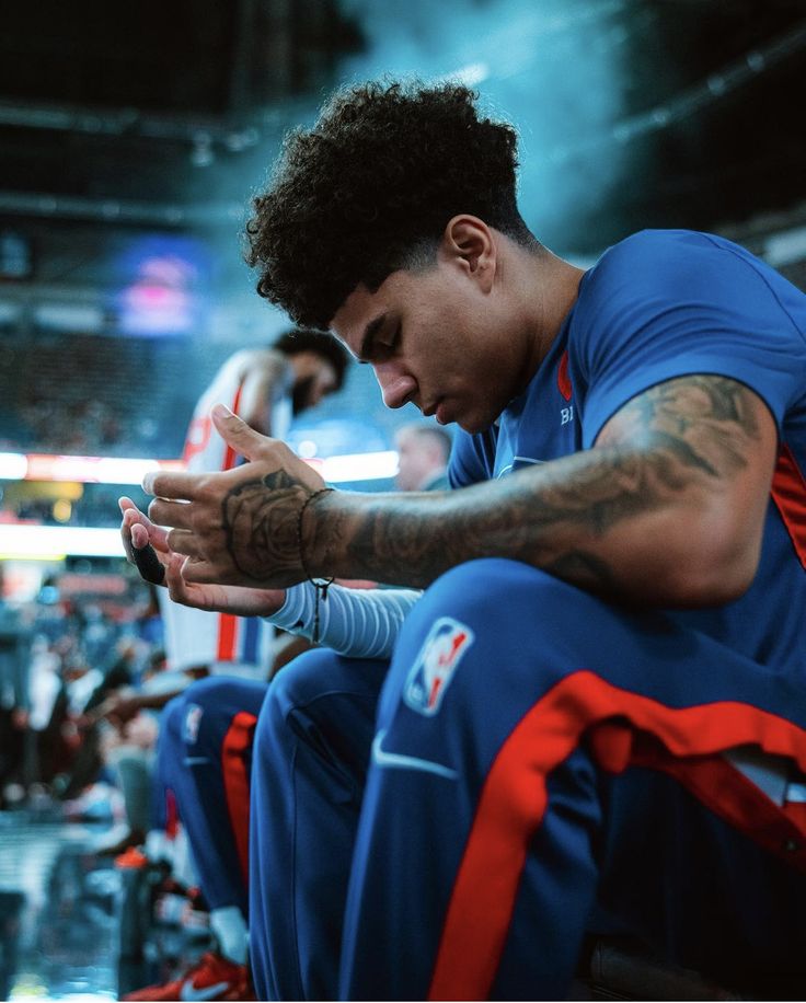 a basketball player sitting on the bench with his hands clasped to his face and other players in the background