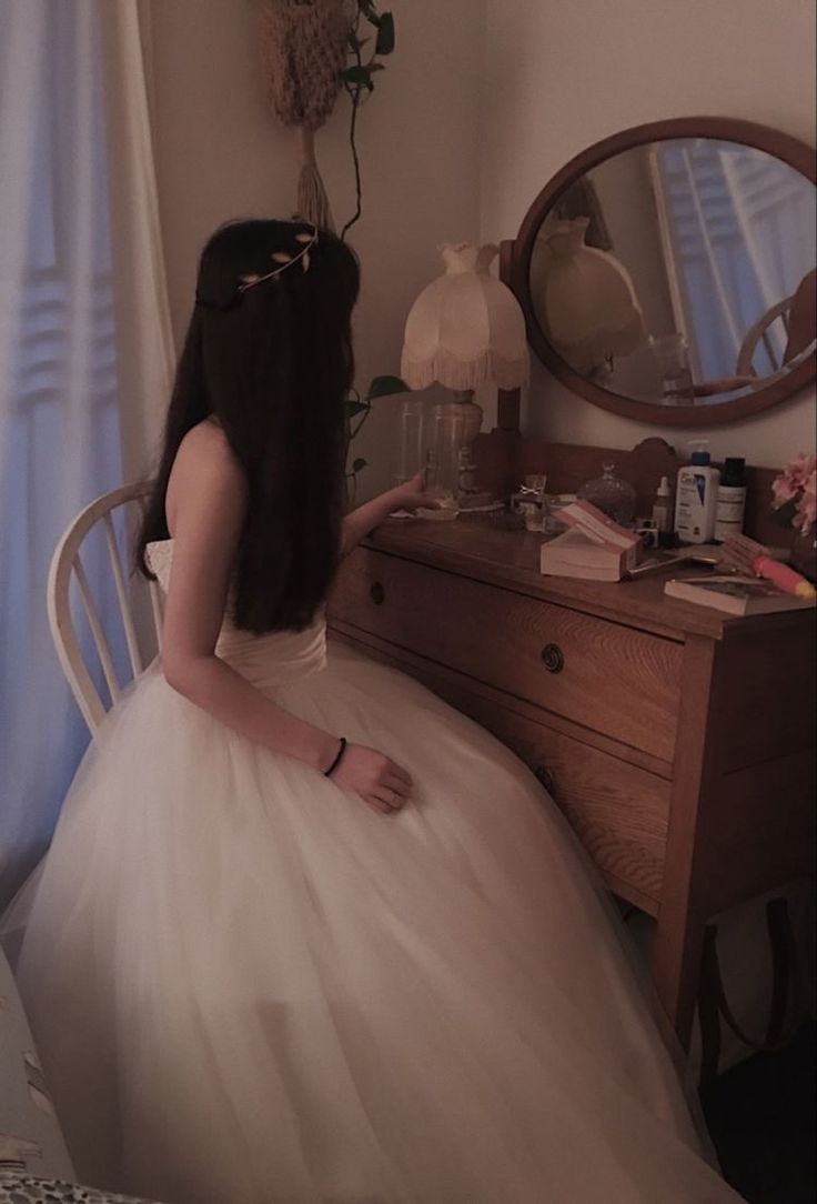 a woman in a white dress is sitting on a chair and looking at the mirror