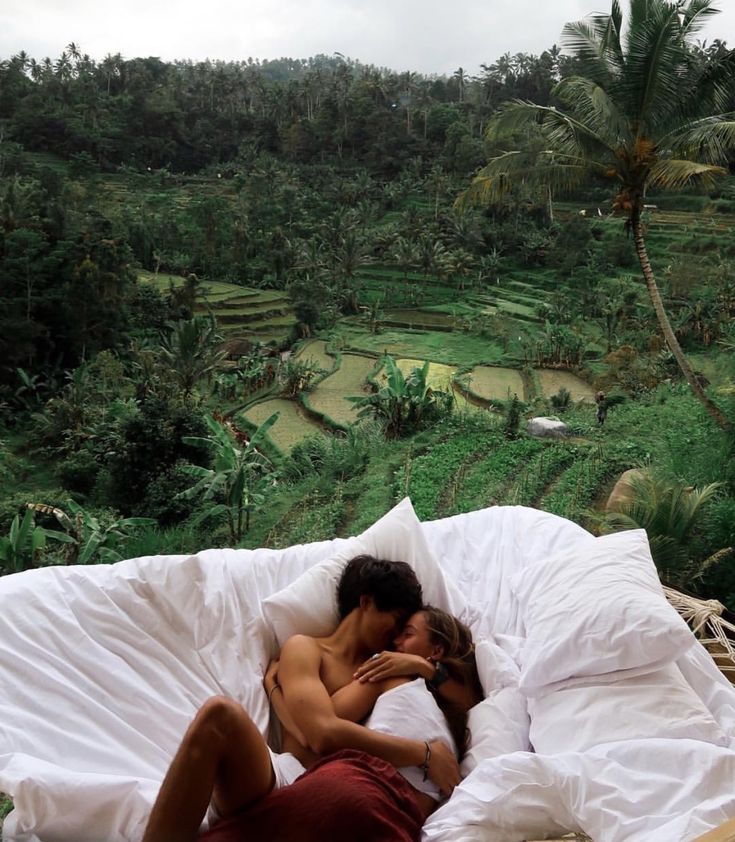 a man and woman laying in bed on top of a lush green hillside with rice fields behind them