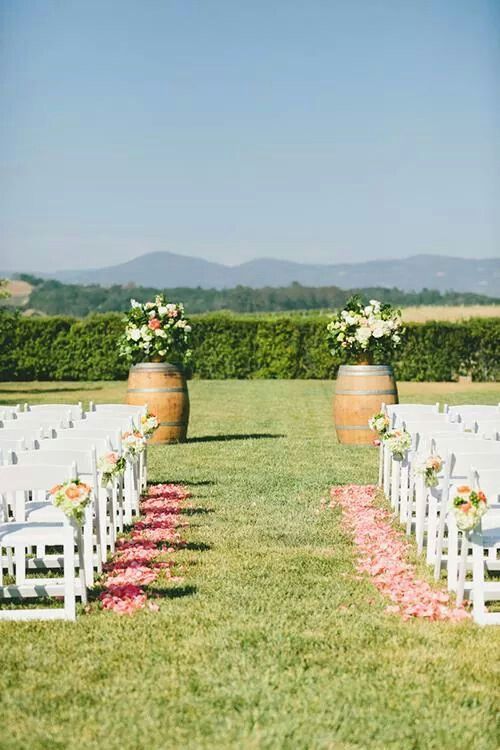 an outdoor ceremony setup with white chairs and flower petals on the aisle, surrounded by wooden barrels