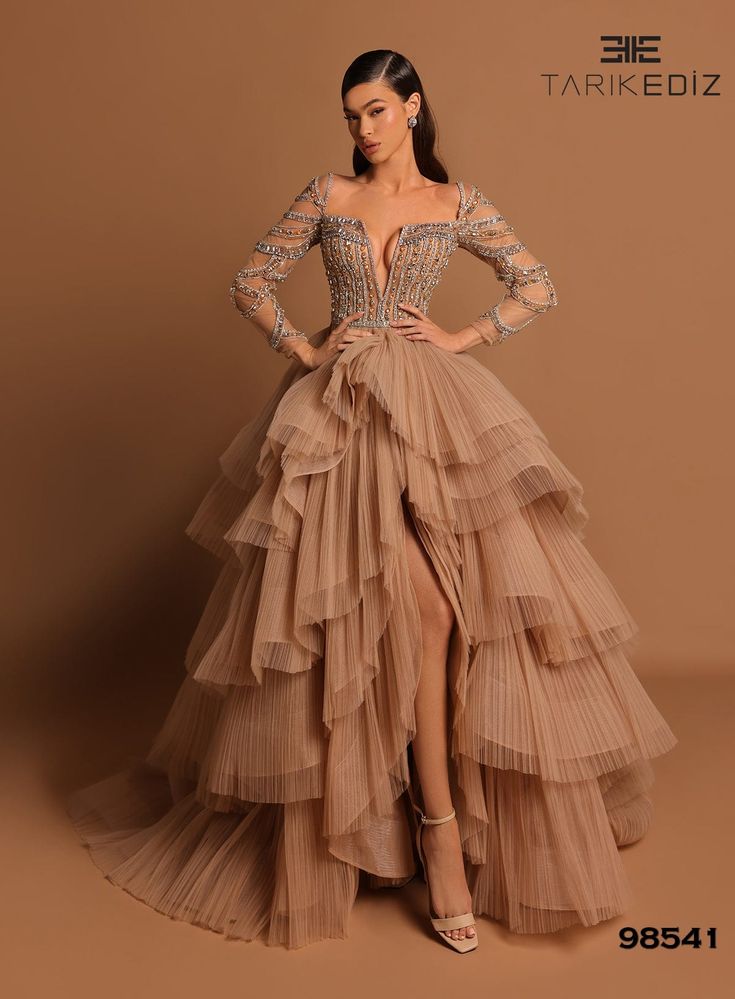 This romantic a-line gown features a full sheer beaded long-sleeved bodice that gives way to a flowy tiered skirt with a sweep train and side slit. The deep v-neck and back zipper closure make it perfect for a special occasion, while the tiered skirt allows you to move freely on the dance floor. A timeless choice for a Tarik Ediz Wedding, Tulle Couture Dress, Gold And White Dresses For Women, Wedding Dress With Textured Skirt, Long Sleeved Tiered Dress, Wedding Guest Ball Gown, Iconic Long Dresses, Full Sleeves Dresses For Women, Haute Couture Blouse