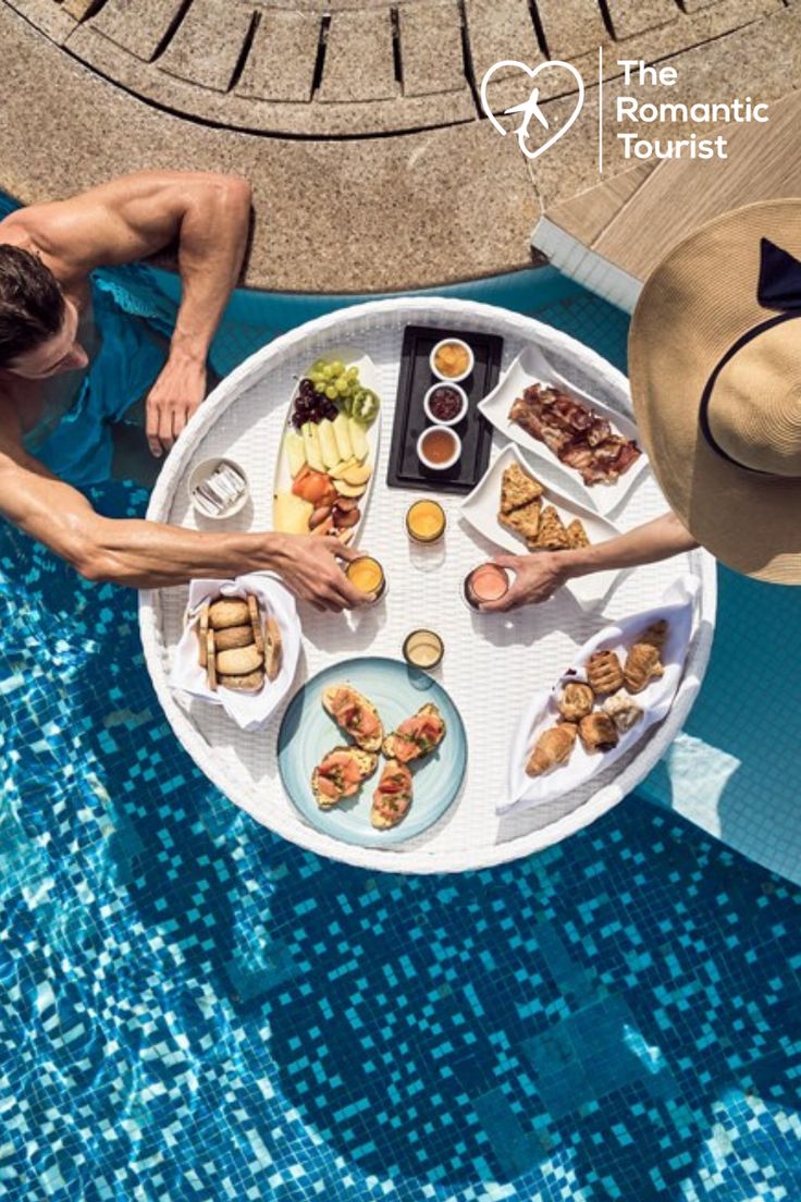 two people sitting at a table with food on it near a swimming pool in the sun