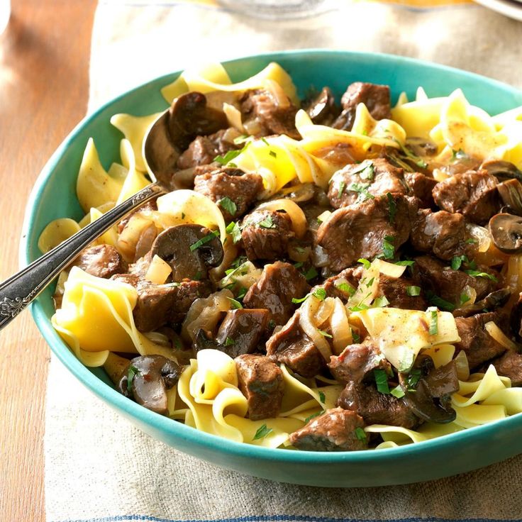 a blue bowl filled with pasta and meat on top of a table next to a fork