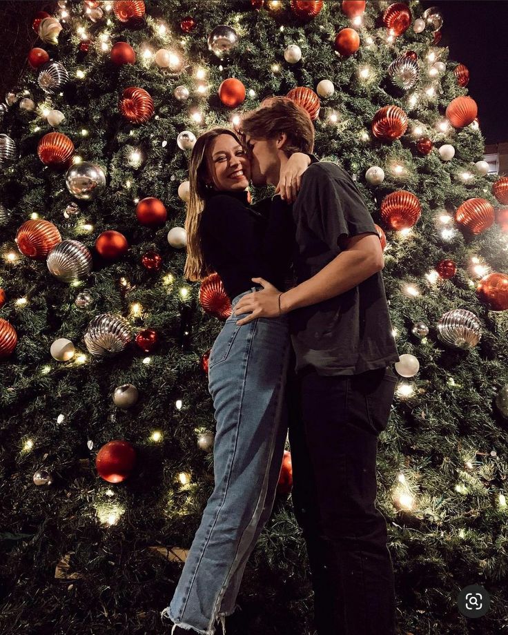 a man and woman hugging in front of a christmas tree with lights all around them