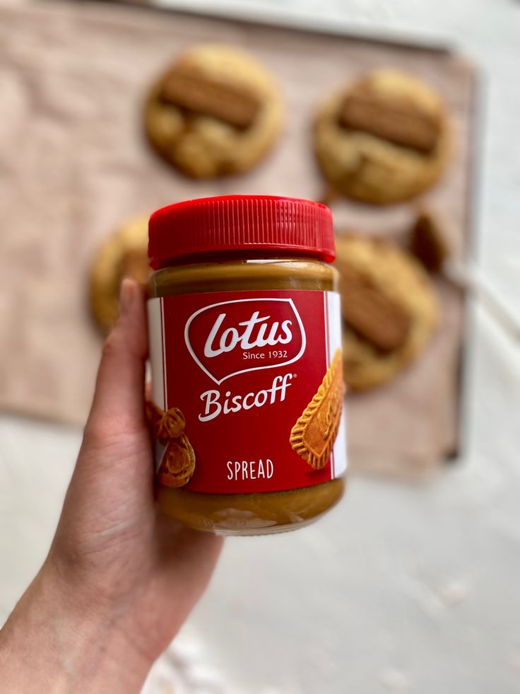 a hand holding a jar of breaded spread in front of cookies on a table