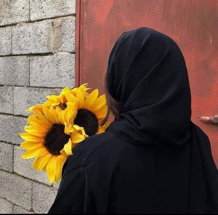 a woman wearing a black hoodie holding a sunflower in front of a brick wall