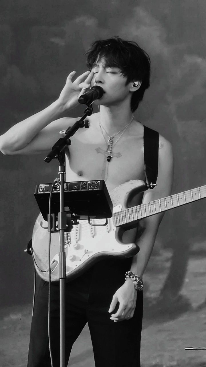 a shirtless man holding a guitar and singing into a microphone while standing in front of a microphone