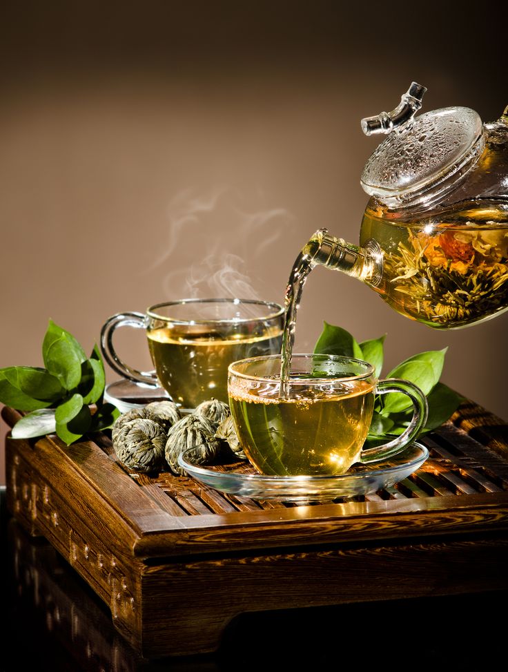 tea being poured into two cups on top of a wooden table with green leaves around it