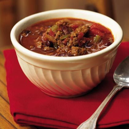 a white bowl filled with chili and meat on top of a red place mat next to a spoon