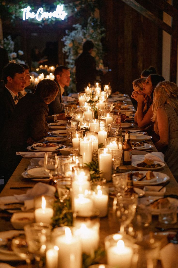 many people are sitting at a long dinner table with candles in front of them and plates on the table