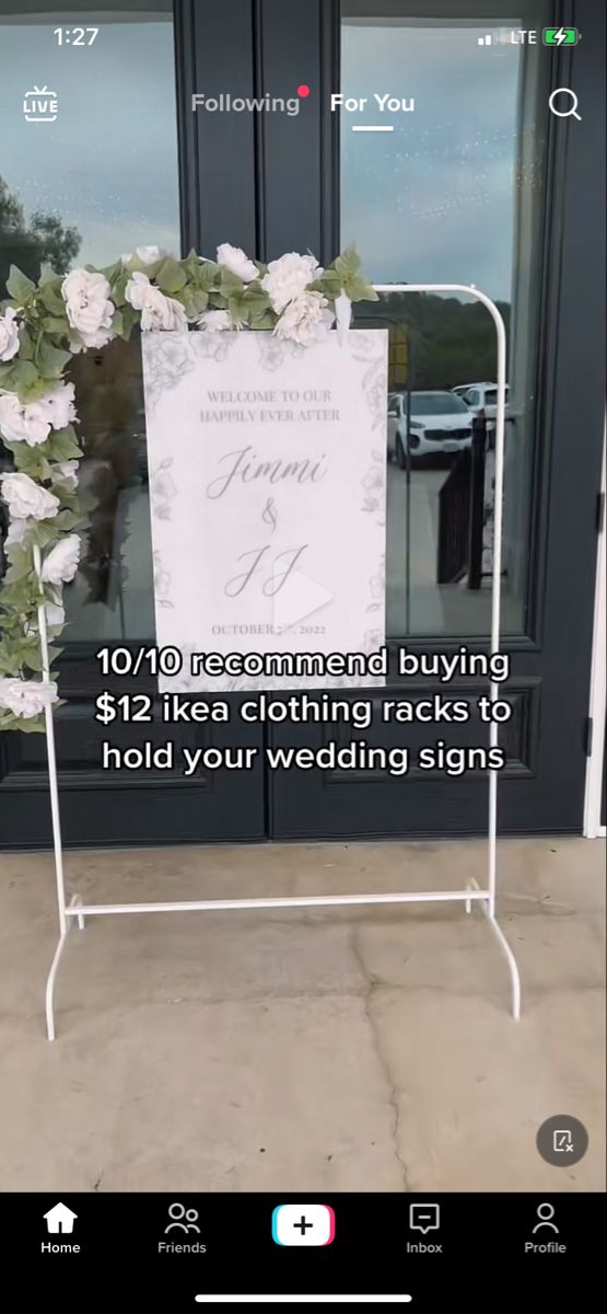 a white sign sitting on top of a sidewalk next to a building with flowers growing out of it