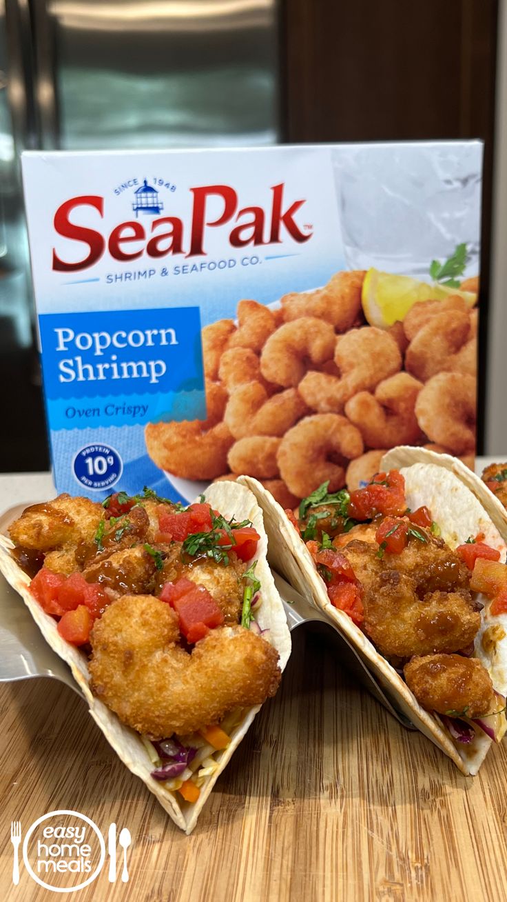 two fish tacos sitting on top of a table next to a box of seapak popcorn shrimp