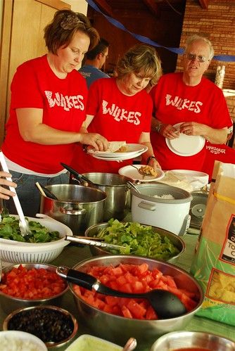 three people in red shirts are preparing food at a table with bowls and spoons