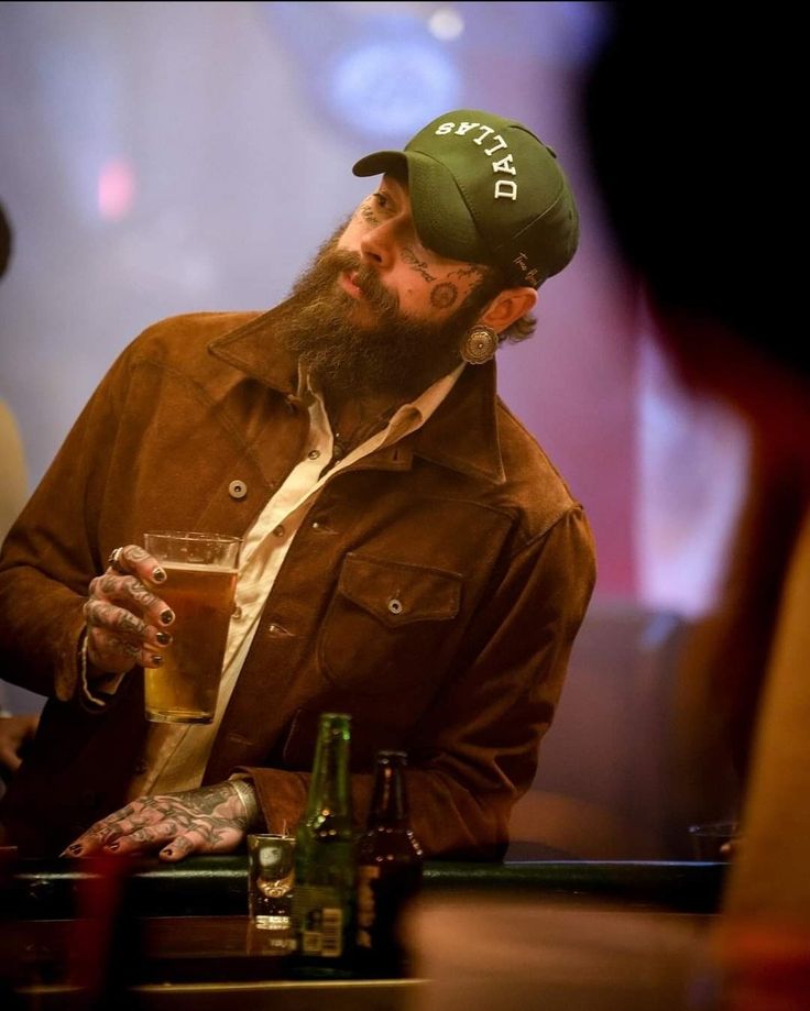 a man with a beard sitting at a bar holding a beer in his right hand