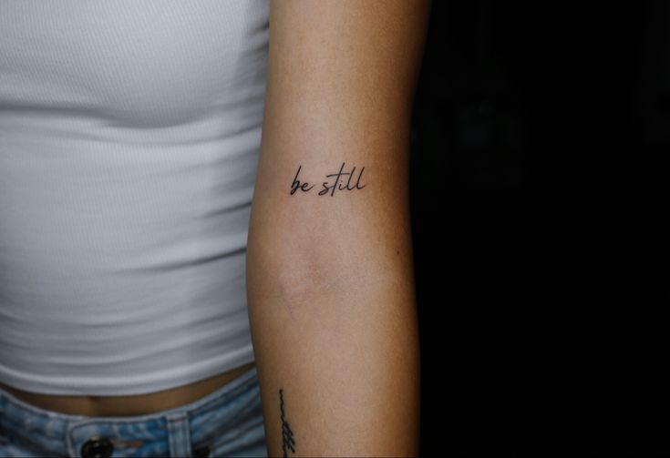 a woman's arm with the words be still tattooed in cursive font