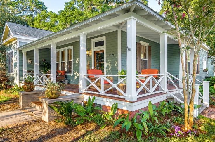 a small blue house with white trim on the front porch and covered porch area, surrounded by greenery