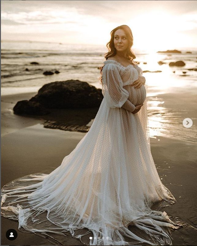 a pregnant woman standing on the beach at sunset wearing a white dress with sheer sleeves