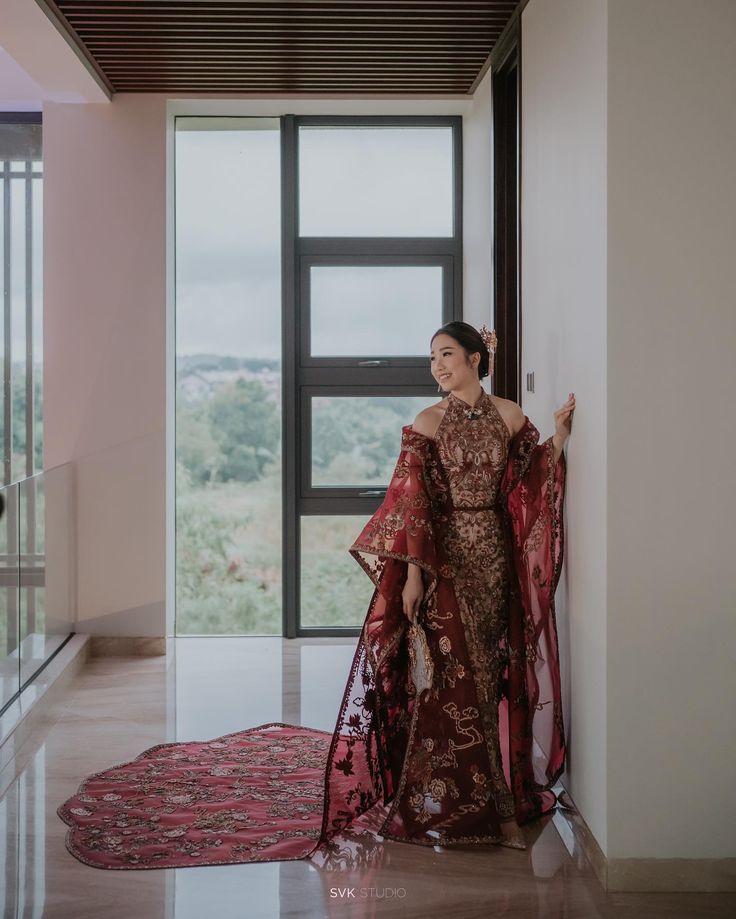 a woman standing in front of a window wearing a red and gold dress with sheer sleeves