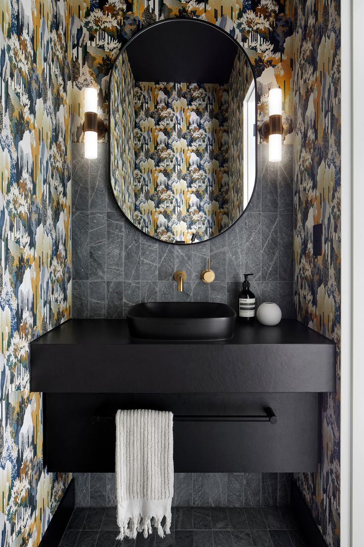 a bathroom with a black counter top and wall paper on the walls, along with a round mirror above it