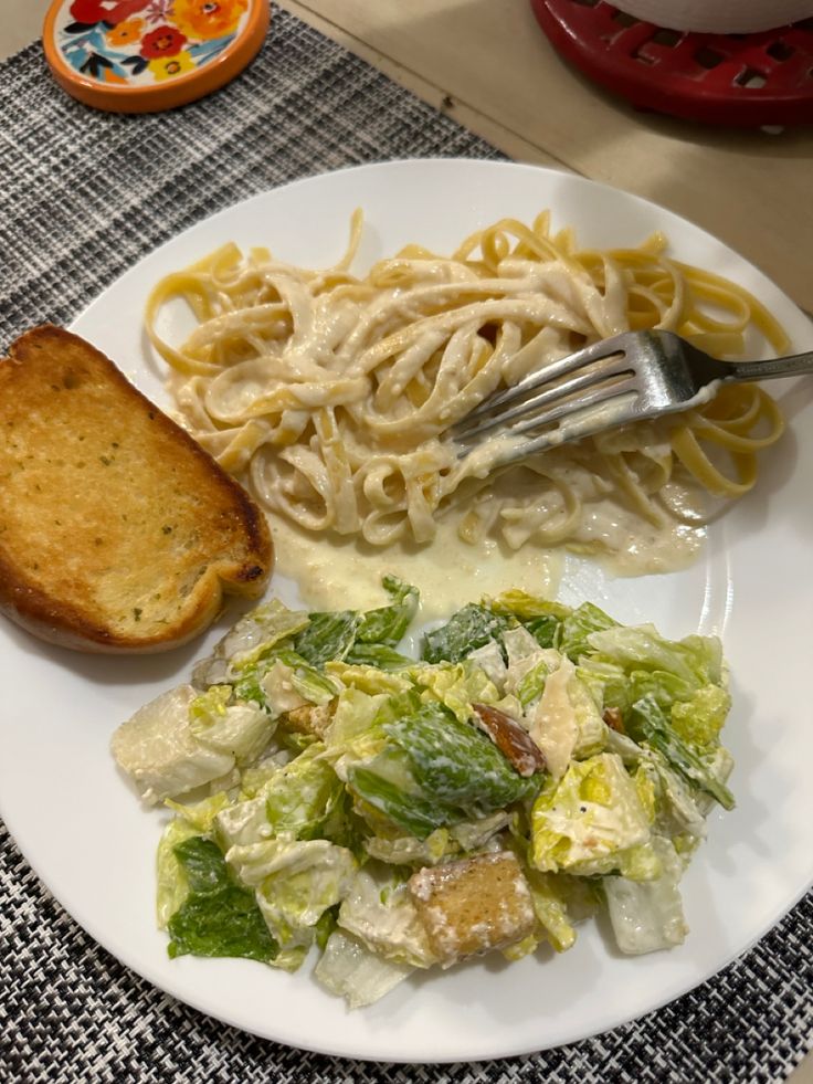 a white plate topped with pasta and salad next to a piece of toasted bread