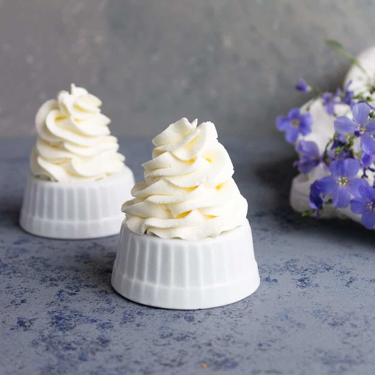 two small white desserts sitting on top of a blue counter next to purple flowers