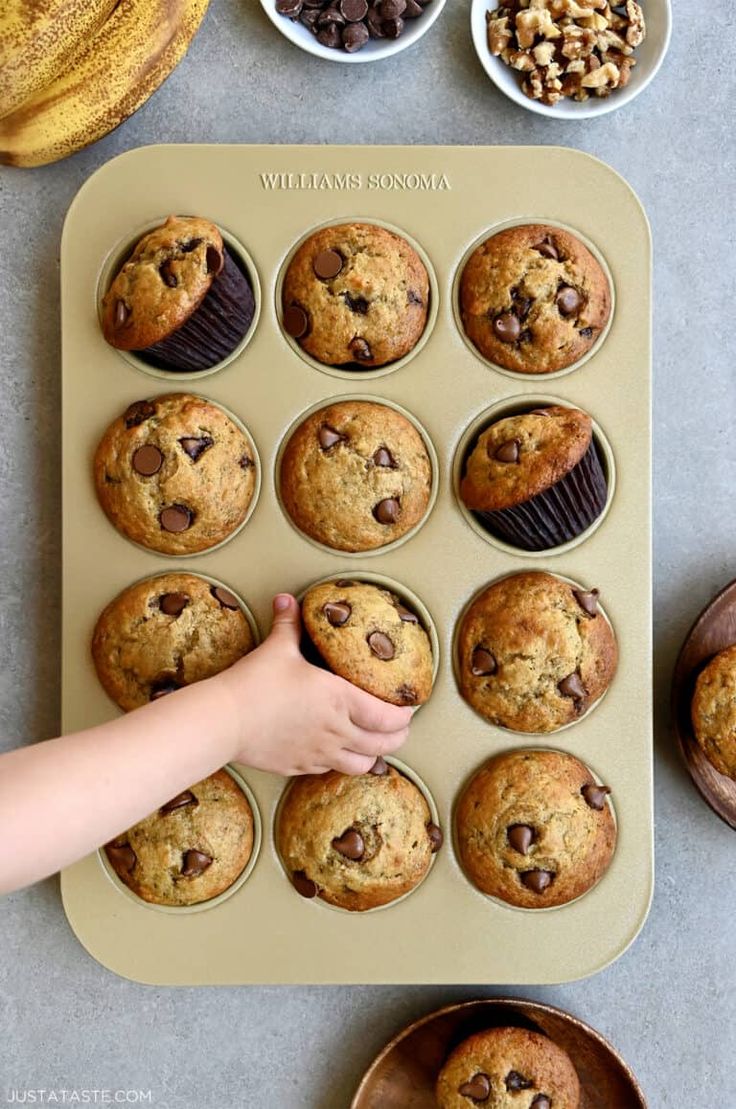 a muffin tray filled with chocolate chip muffins next to bowls of walnuts and bananas