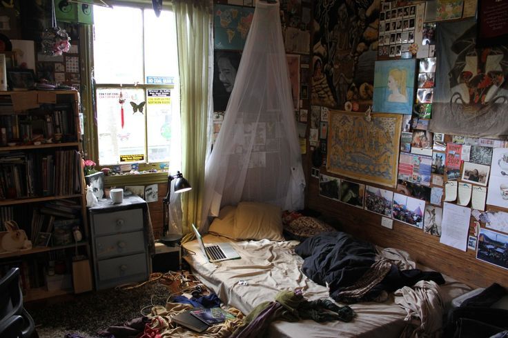 an unmade bed in a cluttered room with posters on the walls and windows