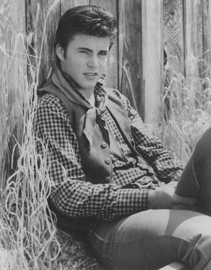 black and white photograph of a young man sitting on the ground in front of a fence