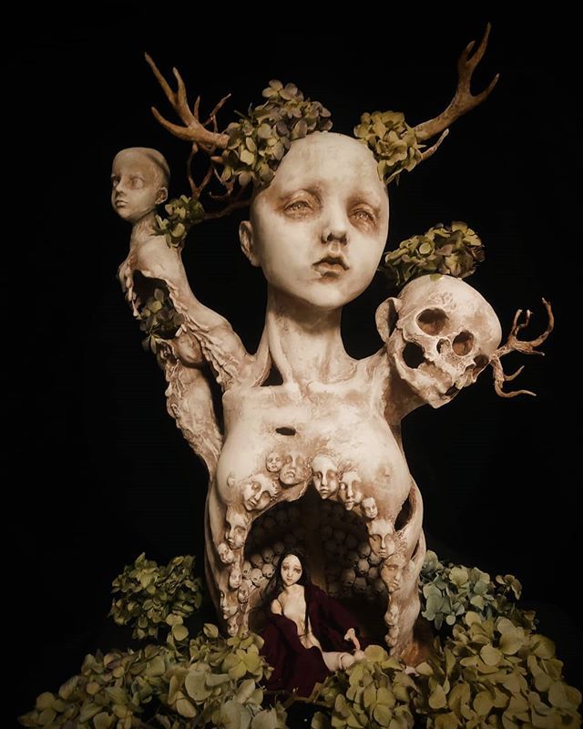 an artistic sculpture with skulls and flowers on the head, surrounded by leaves and branches