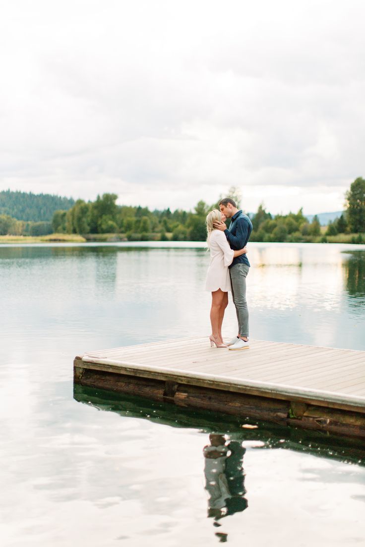 a man and woman kissing on a dock by the water with trees in the background