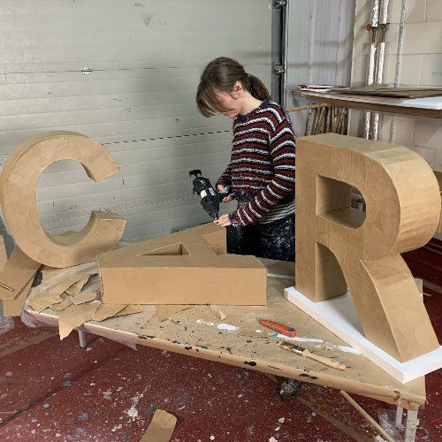 a woman is making letters out of plywood and cardboard with a power drill in her hand