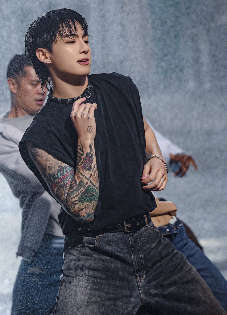 a man with tattoos on his arm standing in front of another man wearing jeans and a t - shirt