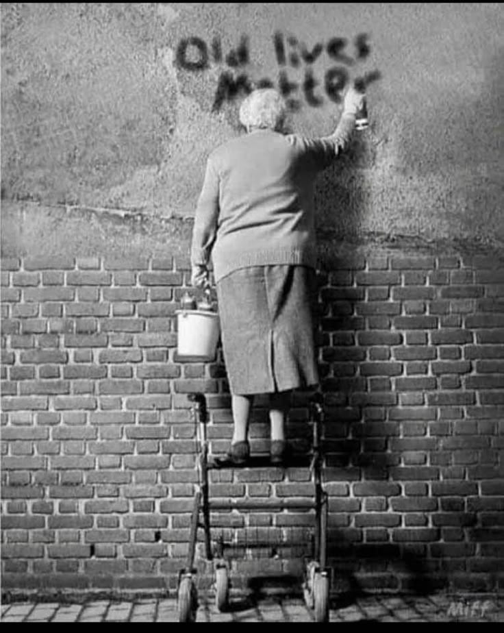 an old woman standing on top of a chair next to a brick wall with graffiti written on it