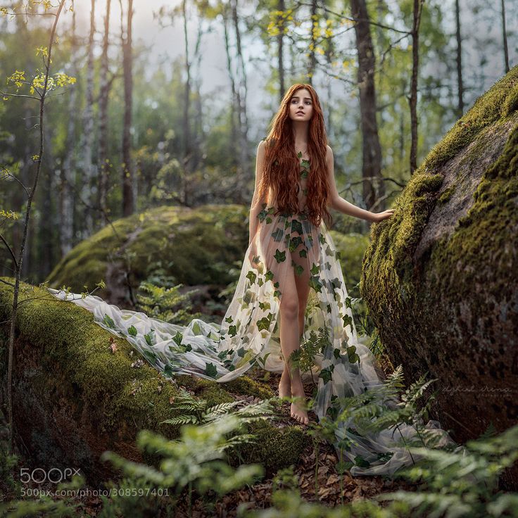 a woman with long red hair standing in the middle of a forest wearing a flowing dress