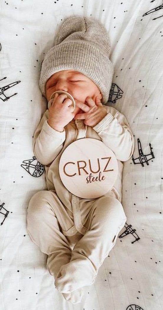 a baby laying on top of a bed wearing a hat and holding a sign that says cruz