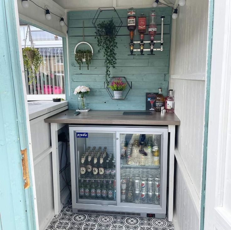 a small bar with lots of bottles and plants on the top shelf, in front of an open door