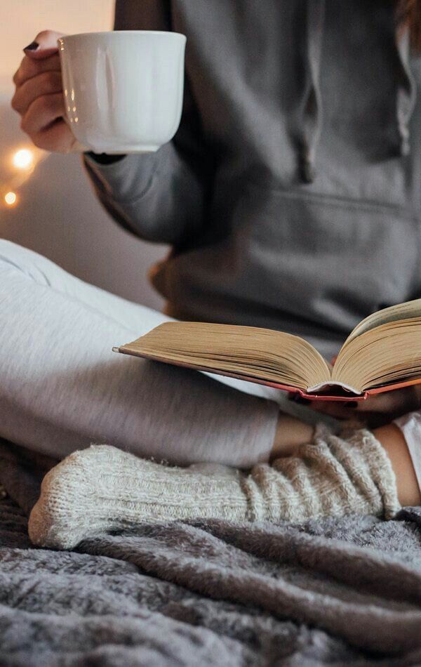 a woman is sitting on the bed while reading a book and holding a coffee cup