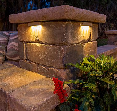 a stone bench with two lights on it next to some plants and flowers in front of the bench