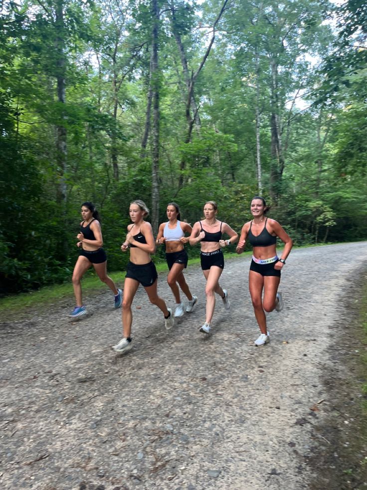 four women running down a dirt road in front of the camera with trees behind them