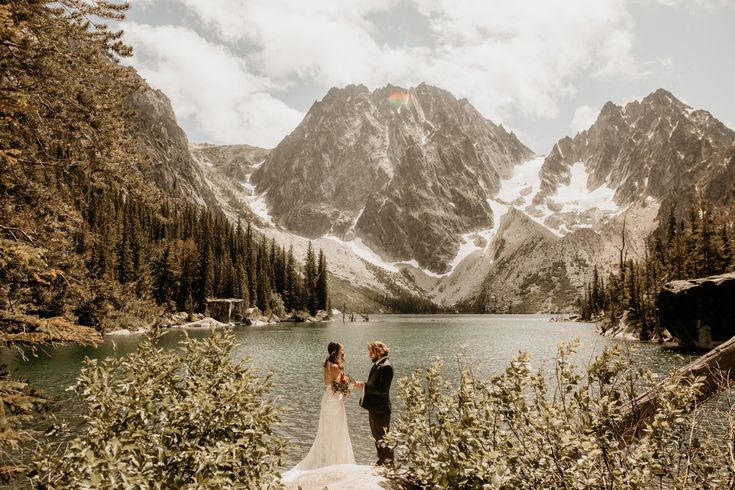 a bride and groom standing in front of a mountain lake with snow on the mountains