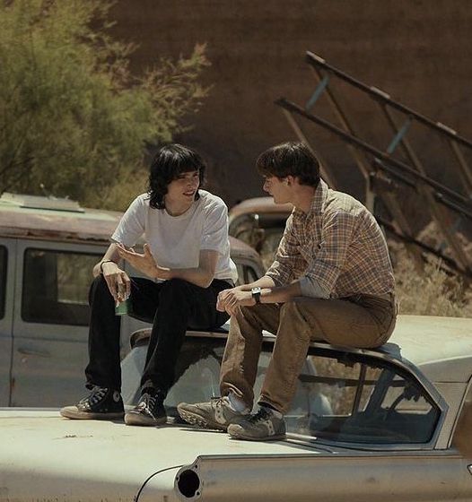 two young men sitting on the hood of an old pickup truck talking to each other