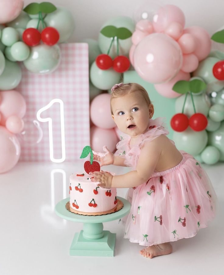 a baby girl in a pink dress standing next to a cake