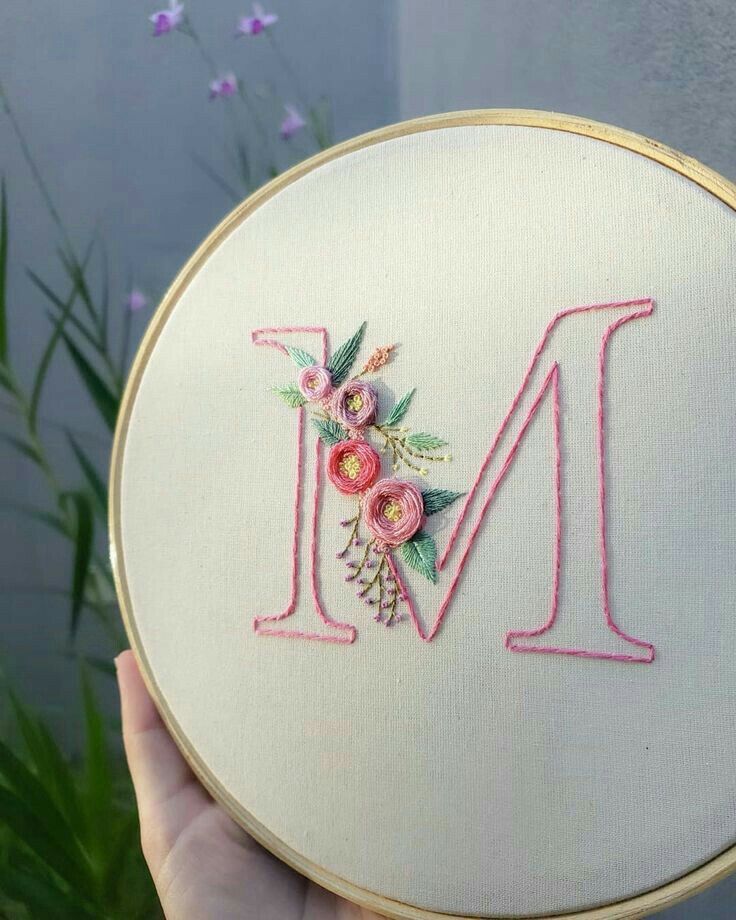 a person holding up a embroideryed letter m in front of some purple and pink flowers