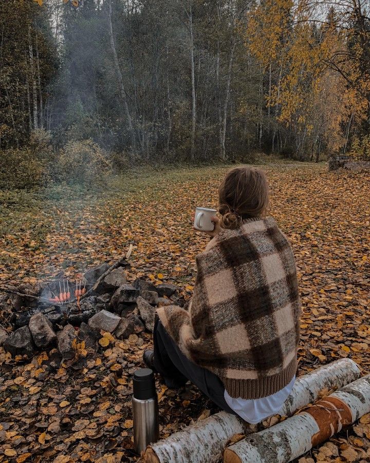 a woman sitting on a log next to a campfire in the woods with her mug