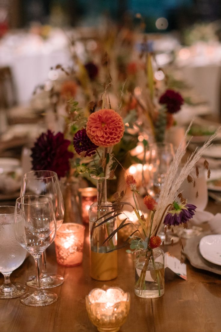 a wooden table topped with lots of vases filled with flowers and candles next to wine glasses