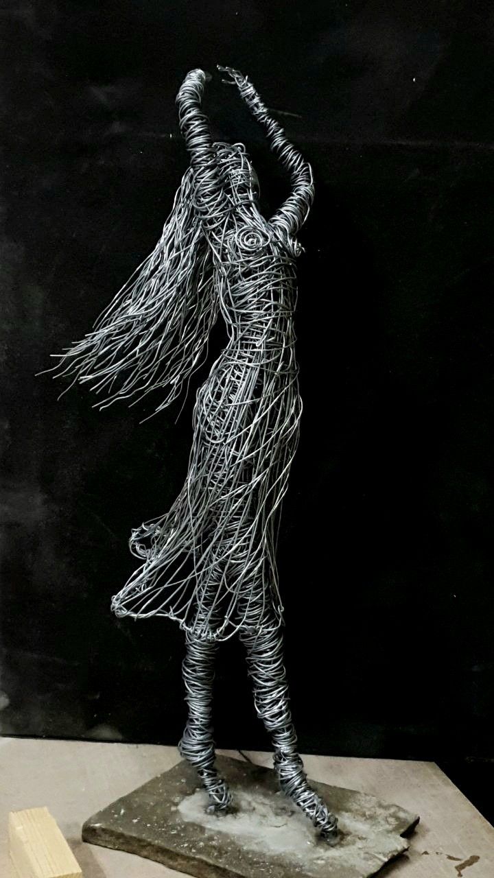 a wire sculpture is standing on top of a block of wood and has long hair blowing in the wind